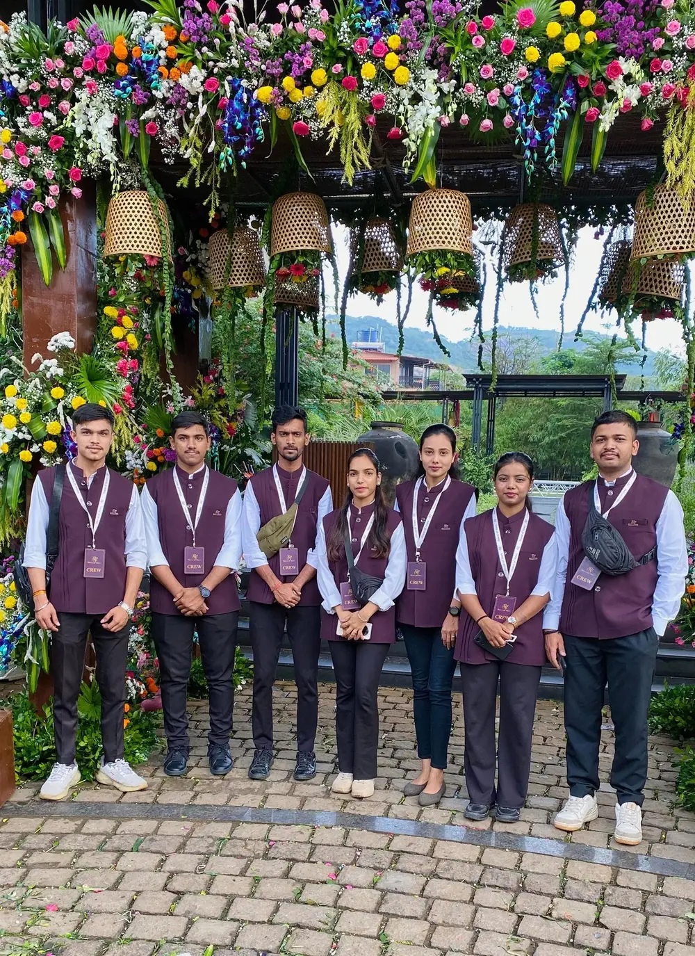 Seven TNP crew members in maroon waistcoats and crew passes under a flower-covered entrance